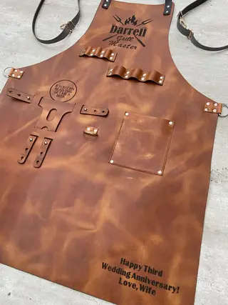 Tan BBQ apron laid flat, showing the pocket, tool loops and beer holder