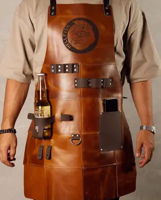 Patterned BBQ apron made of tan leather panels, with a logo engraved on the chest and a bottle in the beer holder
