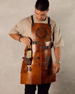 Man wearing a patterned tan leather BBQ apron, with a bottle in the beer holder