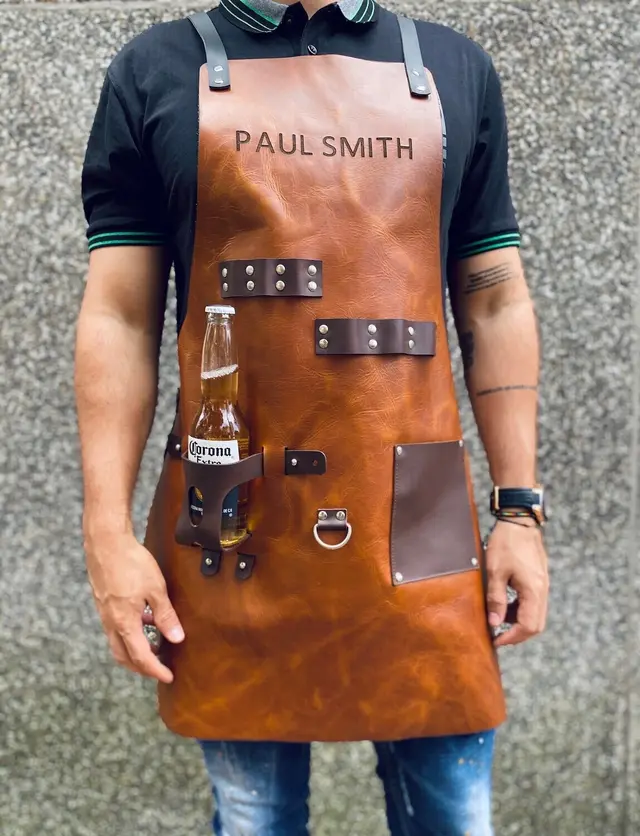 A man wearing a tan leather BBQ apron engraved "Paul Smith", with a beer bottle in the drink holder