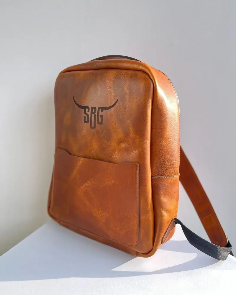 Tan leather minimalist backpack with a front pocket, engraved on the front with a bull's horns and the initials SBG