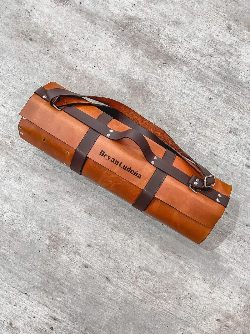 Tan leather knife roll, rolled up, with dark brown straps and shoulder strap, engraved BryanLudeña between the straps