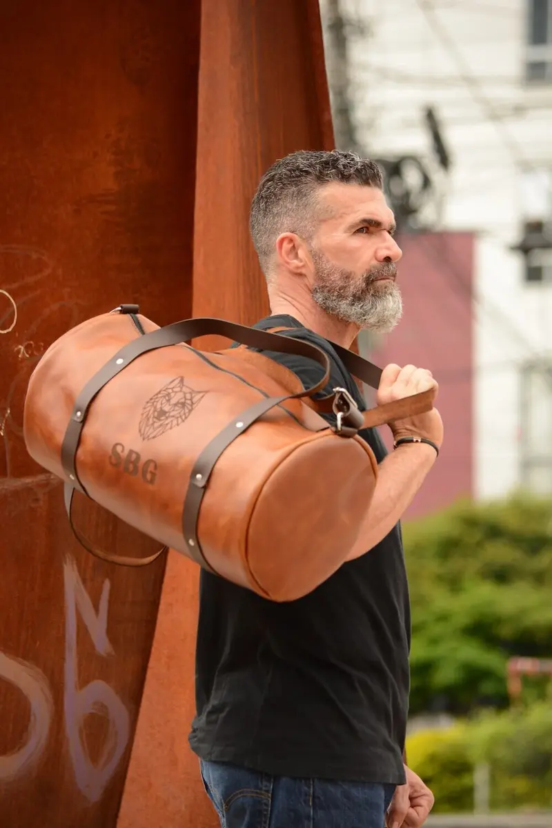Man carrying a tan leather duffle bag with dark brown straps, engraved with a wolf's head and the initials SBG